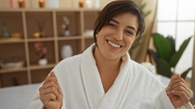 Genç, çekici, İspanyol, esmer saçlı, spa merkezinde beyaz bir bornozla gülümseyen bir kadın.