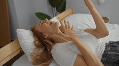 Rahat bir evde, yatakta gülen bir kadın. Sarışın, günlük giyinmiş, güneşli bir yatak odasında giyinmiş. Huzurlu, mutlu bir atmosfer yaratıyor..