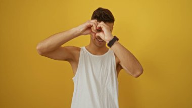 Sarı arka planda gülümseyen genç İspanyol adam kollarını kaldırarak bir kalp şekli yaratıyor, canlı ve dinamik bir portre yakalıyor, pozitiflik ve çekicilik dolu..