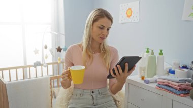 Kadın kahveyi yudumluyor ve bebek odasında tablet kullanıyor. Etrafı bebek malzemeleriyle çevrili. Pencereden içeri güneş ışığı giriyor..