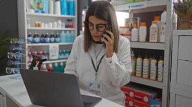 Esmer saçlı ve gözlüklü bir kadın eczacı iyi organize edilmiş bir eczanede dizüstü bilgisayar üzerinde çalışırken telefonla konuşuyor..