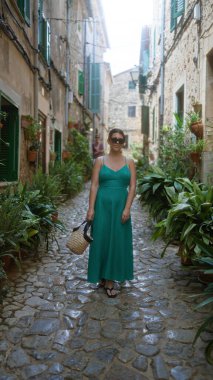 Yeşil elbiseli genç bir kadın Valldemossa, Mallorca, İspanya 'da kaldırımlı caddede yürüyor. Etrafı yemyeşil bitkiler ve geleneksel binalarla çevrili..