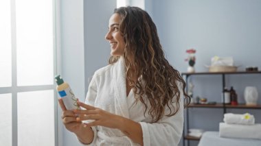 Kaplıca odasında elinde losyonla gezen, beyaz bornoz giyen, gülümseyen ve camdan dışarı bakan esmer saçlı genç bir kadın. Etrafı zenginlik dekoru ve iç mekandaki bitkilerle çevrili.