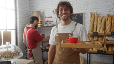 Önlüklü gülümseyen iki erkek fırıncı bir fırında birlikte çalışırken, biri kahve ve kek servis ederken diğeri de raflarda pişmiş yiyecekler hazırlıyor..