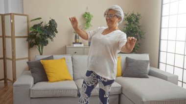Modern mobilya ve bitkilerle dekore edilmiş bir evin oturma odasında dans eden yaşlı kadın neşe ve enerji saçıyor..