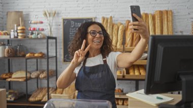 Bir pastanede selfie çeken bir kadın, etrafı ekmek ve pastalarla çevrili, önlük ve gözlük takmış, çeşitli fırınlarla dolu sıcak bir dükkanda eğleniyor..