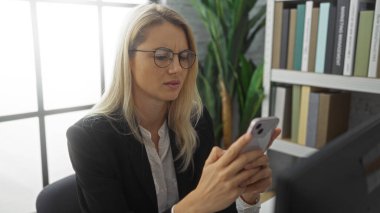 Woman in glasses working on smartphone at office desk with bookshelf in background and potted plant, showing focused and professional expression in corporate environment. clipart