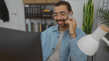Bıyıklı, gözlüklü, modern ofis ortamında telefonla konuşan, arka planda raflar ve bitkiler olan Latin kökenli genç adam.