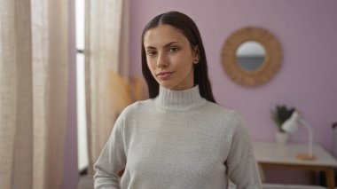 Modern bir oturma odasında mor duvarları olan genç bir İspanyol kadın kameraya özgüvenle bakıyor. Huzurlu bir ev ortamında güzellik ve sükunet yayıyor..