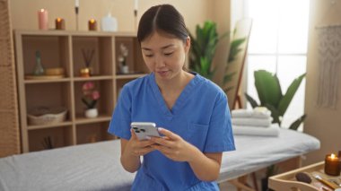 Genç Asyalı bir kadın dinlenme odasında cep telefonu kullanıyor. Arka planda mumlar ve sağlıklı yaşam ortamını gösteren gerekli yağlar var.