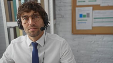 Ofis ortamında kulaklık takan, arka planda raf ve belgelerle profesyonel bir ortamda kendine güvenen, işe odaklı bir atmosfer taşıyan bir adam..