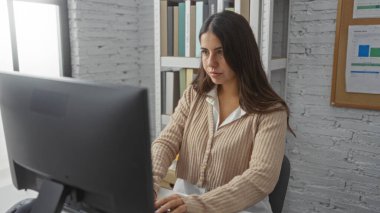 Ofisteki bir masada çalışan bir kadın, bilgisayar ekranına odaklanmış, modern bir çalışma alanındaki kitaplar ve belgelerle çevrili..