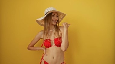 Kırmızı bikinili ve geniş kenarlı şapkalı bir kadın canlı sarı bir arkaplanı işaret ediyor.