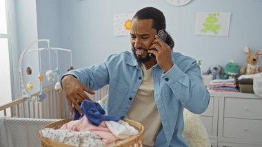 Parlak bir çocuk odasında beşikte bebek kıyafetlerini düzenlerken telefonla konuşan bir adam, odak ve endişeyi kapalı alanlarda ifade ediyor..