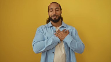 Sakallı, mavi tişörtlü, sarı arka planda gözleri kapalı gülümseyen barış ve sakinlik saçan ve elleri göğsünde şükranlarını ifade eden bir adam..