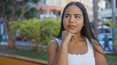 Şehir merkezindeki ağaçların arka planında, uzun esmer saçlı, düşünceli bir ifadeyle genç ve çekici bir kadını sergiliyor..