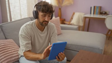 Rahat bir oturma odasında otururken tablet kullanan genç adam rahat bir ev ortamı, kanepe, yumuşak yastıklar ve arka planda bir masa öneriyor..