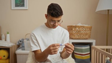 Bebek odasındaki genç adam hamilelik testini inceliyor. Etrafı rahat dekor ve beşiklerle çevrili. Ebeveynlik anlarında beklenti ve sıcaklık gösteriyor.