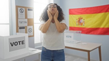 Mature woman feeling stressed in an electoral college room with a spanish flag and a voting booth clipart