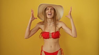 Kırmızı bikinili ve güneş şapkalı güzel sarışın bir kadın izole edilmiş sarı arka plan duvarına karşı coşkuyla poz veriyor..