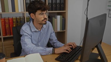 Ofis ortamında sakallı ve gözlüklü, kitaplarla ve klasörlerle çevrili bir bilgisayarda çalışan İspanyol bir adam görevlerine odaklandı..