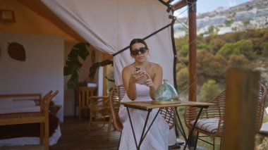 Güneş gözlüğü takan, beyaz elbiseli, genç ve güzel bir İspanyol kadın Afrika 'da bir kamp çadırında akıllı telefonunu kullanarak bir masada oturuyor..