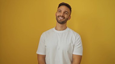 Sakallı genç bir İspanyol, portrede izole edilmiş sarı arka plan duvarına gülümsüyor. Beyaz gömlek giyiyor..