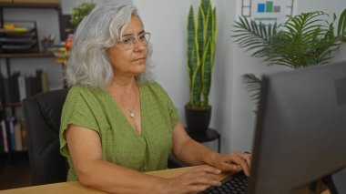 Ofiste bilgisayar başında çalışan yaşlı bir kadın. Kapalı mekanlar, kitaplıklar ve grafik grafikleriyle.