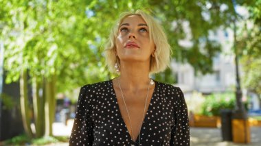 Şehir parkında kısa sarı saçlı, güneş ışığı altında noktalı elbise giyen bir kadın.