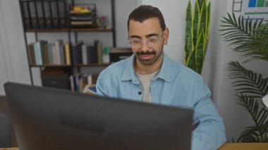 Ofiste çalışan İspanyol bıyıklı bir adam, bilgisayara odaklanmış, kitaplıklar, kapalı mekanlar ve ofis dekorlarıyla çevrili..