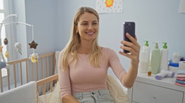 Kadın rahat bir yatak odasında bebek beşiği ile selfie çekerken gülümsüyor. Rahatlamış bir atmosfer ve etrafa saçılmış kişisel bakım ürünleri sergiliyor..