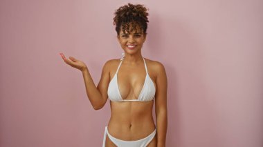 Kadın elini uzatmış, beyaz bir bikini giymiş pembe bir arka planda güzel bir gülümseme, kendine güvenen bir poz ve kıvırcık saç stili sergiliyor..