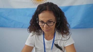 Woman working indoors with glasses and headset, argentinian flag behind her, at an electoral college room. clipart