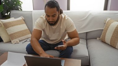 Konforlu oturma odası ortamında dizüstü bilgisayar ve akıllı telefon kullanan, odaklanmış ifadesi ve rahat duruşu olan, günlük kıyafetler giyen ve nötr tonlarla çevrili bir adam..