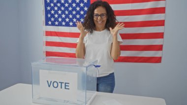 Kapalı bir Amerikan seçim salonunda arka planda Birleşik Devletler bayrağı ve önünde bir oy sandığıyla kutlama yapan olgun bir kadın..