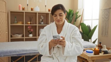 Beyaz bir bornozla kaplıcada dinlenen ve akıllı telefon kullanırken gülümseyen bir kadın. Etrafı mumlarla çevrili..