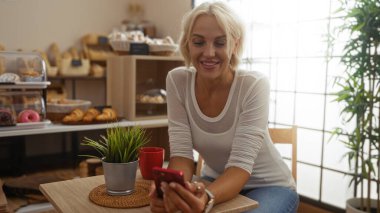 Pastanede gülümseyen, arka planda pastalar olan bir kadın, bir bitkiyle masada otururken elinde bir telefonla, beyaz bir gömlek giyiyor.