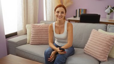 Çekici kızıl saçlı kadın rahat bir oturma odasında kanepede oturmuş elinde uzaktan kumandayla gülümsüyor.