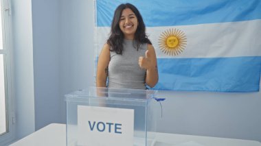 Arjantin 'de kapalı bir üniversite odasında oy verme kabini ve Arjantin bayrağıyla gülümseyen ve onaylayan genç, çekici esmer bir kadın..