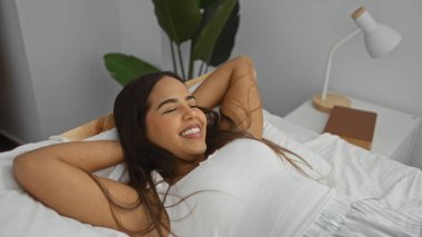 Modern bir yatak odasında bir yatakta dinlenen, minimalist bir dekora gülümseyen bir kadın, gün içinde ev ortamında rahatlık ve sükuneti yakalar..