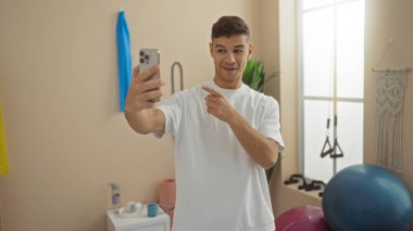 Spor salonundaki genç adam akıllı telefonuyla selfie çekiyor, gülümsüyor ve işaret ediyor, yoga minderleri ve fitness topları gibi egzersiz ekipmanlarıyla çevrili, aktif bir yaşam tarzını somutlaştırıyor..