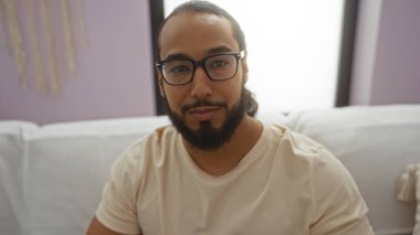 Gözlüklü ve sakallı genç adam modern bir apartman dairesinde oturuyor. Yumuşak ışıklandırma ve ince dekorla rahat ve çağdaş bir atmosfer yaratıyor..