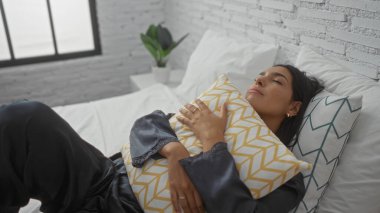 Genç bir kadın, rahat bir yatak odasında, rahat bir şekilde yatağında elinde desenli bir yastıkla uzanıyor..