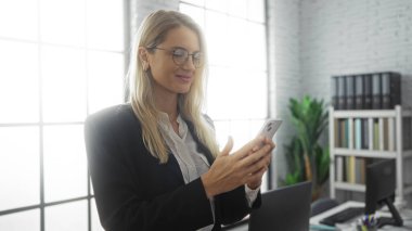 Ofisteki kadın, modern mobilyalar ve parlak iç mekan ile çevrili gözlük takmış akıllı telefona bakarken gülümsüyor..