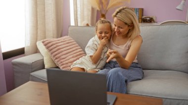 Kadın ve kız rahat bir oturma odasında kanepede oturuyor, gülümsüyor ve açık bir dizüstü bilgisayarla iletişim kuruyor, sıcak bir aile ortamı yaratıyor..