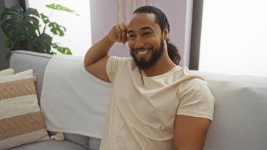 Oturma odasında sırıtan sakallı genç bir adam. Kanepede oturmuş yastık dekoru ve pencereden süzülen rahatlatıcı ortam ışığıyla..