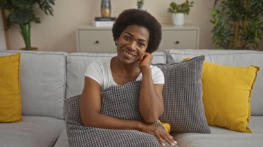 Güzel Afro-Amerikalı kadın, kıvırcık saçlı, sarı yastıklı bir kanepede oturuyor, iyi aydınlatılmış bir oturma odasında, kameraya gülümsüyor, elinde gri bir yastık tutarken.