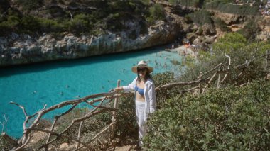 Genç bir kadın Mallorca, İspanya 'daki Cala del Moro manzarasının keyfini çıkarıyor, berrak turkuaz suların ve güneşli gökyüzünün altındaki doğal kayalıkların yanında duruyor..