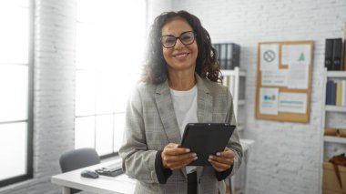 Modern ofiste elinde tabletle gülümseyen bir kadın, iş kıyafetleri ve parlak iç mekanıyla profesyonel ve üretken bir atmosfer ve neşeli bir ortam izlenimi veriyor..