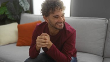 Kıvırcık saçlı genç adam içeride gülümsüyor. Rahat atmosferi ve doğal ışığı olan bir oturma odasında öylesine oturuyor.